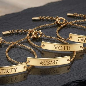 May include: Gold-toned bracelets with engraved messages: "LIBERTY", "RESIST", "VOTE", and "FREEDOM". Each bracelet features a delicate chain and a rectangular bar with the word engraved in a serif font. The bracelets are displayed on a dark, textured surface.