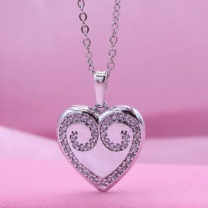 May include: A silver heart-shaped pendant necklace. The pendant is encrusted with small, clear crystals around the edges and features a decorative swirl design. The necklace hangs on a delicate silver chain against a pink background.