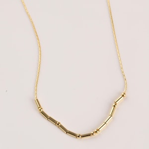 May include: A delicate gold necklace featuring small, cylindrical and spherical beads. The beads are strung on a fine gold chain. The necklace is displayed against a plain white background.