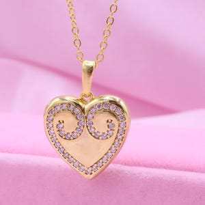 Puede incluir: Un collar con colgante de corazón dorado. El colgante en forma de corazón está adornado con pequeñas piedras preciosas transparentes en los bordes y dos diseños en espiral. La cadena del collar también es dorada, sobre un fondo rosa suave.