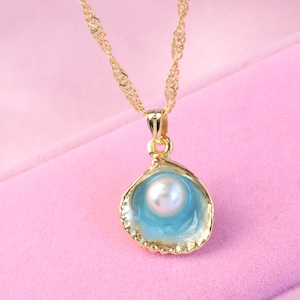 May include: A gold-tone twisted chain necklace featuring a pendant shaped like a seashell. The interior of the shell is painted a bright sky blue and contains a single, round, white pearl centered within the shell's cavity.