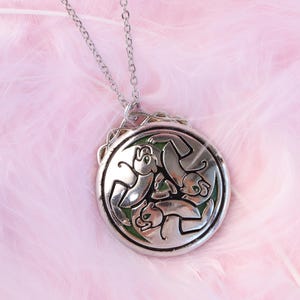 May include: A silver-toned circular pendant necklace featuring an intricate relief design of three stylized animals arranged in a rotating pattern. The metalwork is set against a dark green background, and the pendant hangs from a thin silver chain.