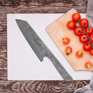 May include: A Damascus steel chef's knife with a textured blade and handle rests on a white cutting board. Cherry tomatoes are on a wooden cutting board next to the knife. The knife has a unique pattern.