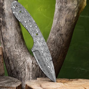 May include: A Damascus steel knife blade with a unique swirling pattern. The blade has a pointed tip and a curved edge. The handle area has three holes. The knife is displayed against a natural wood background with a green backdrop.