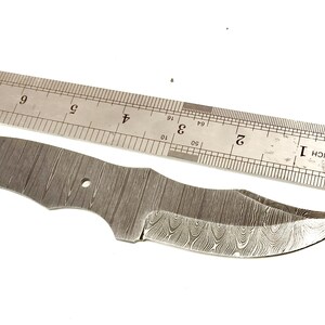 Pu&ograve; includere: Una lama di coltello in acciaio Damasco con un bordo curvo e un motivo strutturato. Un righello di metallo &egrave; posto sopra la lama. La lama &egrave; lunga circa 18 cm.