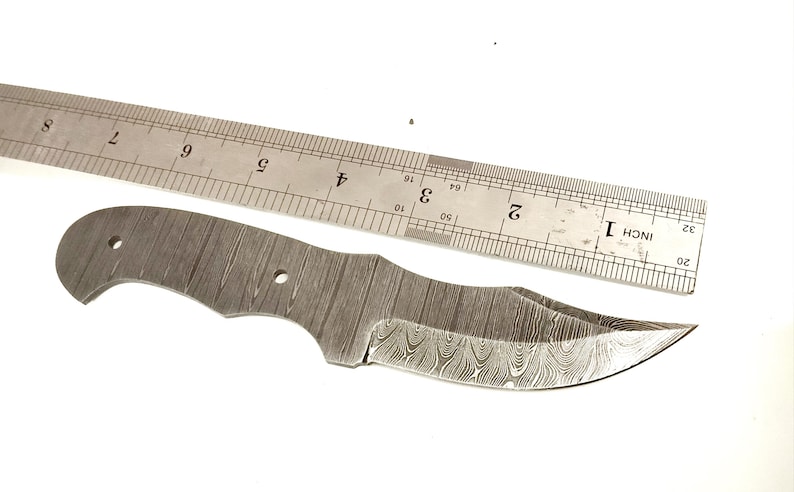 Pu&ograve; includere: Una lama di coltello in acciaio Damasco con un bordo curvo e un motivo strutturato. Un righello di metallo &egrave; posto sopra la lama. La lama &egrave; lunga circa 18 cm.