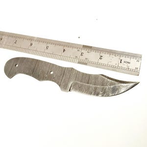 Pu&ograve; includere: Una lama di coltello in acciaio Damasco con un bordo curvo e una superficie a motivi. Un righello di metallo &egrave; posto accanto alla lama. Il righello misura fino a 30 cm.