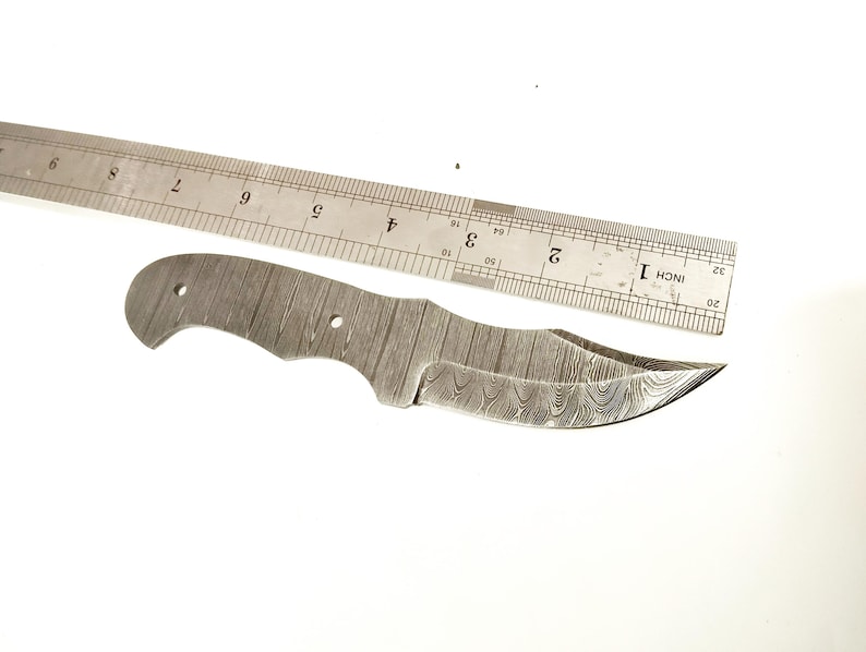 Pu&ograve; includere: Una lama di coltello in acciaio Damasco con un bordo curvo e una superficie a motivi. Un righello di metallo &egrave; posto accanto alla lama. Il righello misura fino a 30 cm.