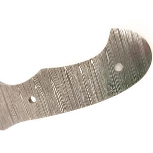 Pu&ograve; includere: Primo piano di una lama di coltello in acciaio Damasco. La lama ha una forma curva con un motivo a venature del legno e due fori per il fissaggio. Il metallo &egrave; di colore argento e grigio.