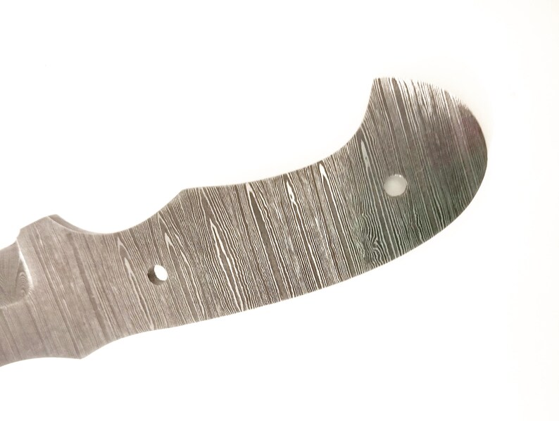 Pu&ograve; includere: Primo piano di una lama di coltello in acciaio Damasco. La lama ha una forma curva con un motivo a venature del legno e due fori per il fissaggio. Il metallo &egrave; di colore argento e grigio.