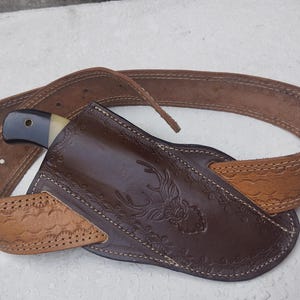 May include: A brown leather belt with a decorative pattern and a brown leather sheath for a knife. The knife has a black handle and a white blade.