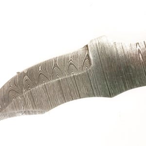 Pu&ograve; includere: Primo piano di una lama di coltello parzialmente rifinita con un caratteristico motivo a venature del legno. La lama ha un bordo curvo e un foro vicino all'impugnatura. Il metallo ha un aspetto scuro e strutturato.