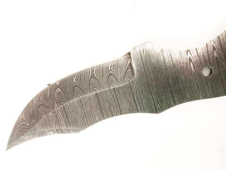 Pu&ograve; includere: Primo piano di una lama di coltello parzialmente rifinita con un caratteristico motivo a venature del legno. La lama ha un bordo curvo e un foro vicino all'impugnatura. Il metallo ha un aspetto scuro e strutturato.