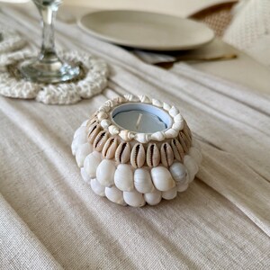 Peut inclure: Un bougeoir décoratif rond, orné de coquillages blancs et beiges. Il contient une petite bougie. Les coquillages sont disposés en couches, créant une surface texturée. Le bougeoir est posé sur une nappe beige.
