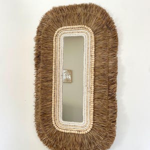 Può includere: Uno specchio da parete rettangolare con un design naturale e bohémien. Lo specchio è incorniciato con un bordo di materiale intrecciato marrone chiaro, corda color crema e piccole perline di legno. La cornice esterna ha un bordo sfrangiato.