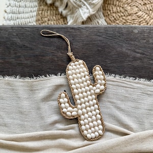 Puede incluir: Decoración de pared con forma de cactus. El cactus está hecho de pequeñas cuentas redondas blancas y delineado con un material similar a una cuerda. Tiene un lazo en la parte superior para colgar. El fondo incluye una superficie de madera y una tela beige.