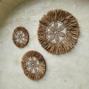 Op de afbeelding: Drie ronde wanddecoraties gemaakt van natuurlijke geweven vezels. Elk stuk heeft een centraal ontwerp van witte kralen en een bloemachtig patroon. De grootste is ongeveer 50 cm in diameter, de andere zijn kleiner.