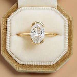 May include: A gold ring with an oval-cut diamond, set in a bezel setting. The ring is displayed in an open, octagonal, velvet ring box with a cream-colored interior. The box is a muted gold color.
