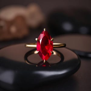 Op de afbeelding: Een gouden ring met een grote, marquise-geslepen, dieprode edelsteen. De edelsteen wordt vastgehouden door vier klauwen en de band is van effen goud. De ring wordt getoond op een donker, reflecterend oppervlak.