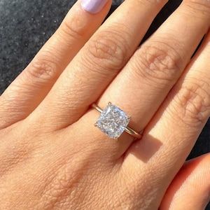May include: A gold ring with a large, square-cut diamond. The diamond is clear and sparkling, set on a thin gold band. The ring is shown on a hand with light skin and painted nails.