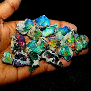 Puede incluir: Una colección de piedras preciosas de ópalo en bruto en varios tonos de verde, azul y rojo. Los ópalos tienen un brillo oscuro e iridiscente y tienen una forma irregular.