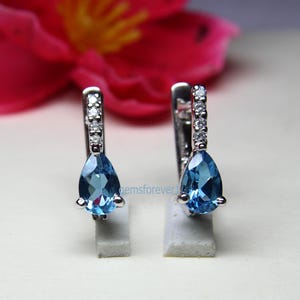 Peut inclure: Une paire de boucles d'oreilles pendantes argentées. Chaque boucle d'oreille présente une pierre précieuse bleue clair en forme de poire et une rangée de petites pierres claires. Les boucles d'oreilles sont présentées sur de petits supports blancs.