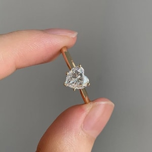 May include: A gold ring featuring a heart-shaped diamond. The ring's band is a thin, polished gold, and the diamond is held in place by small prongs. The diamond sparkles with multiple facets, reflecting light.