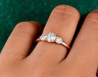 Può includere: Un anello d'oro con tre pietre preziose rotonde e trasparenti. La pietra centrale è più grande delle due laterali. L'anello è mostrato su un dito su uno sfondo verde scuro.