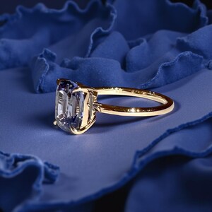 May include: A gold ring with a large, rectangular, light blue gemstone. The ring is set against a deep blue, textured background, creating a striking contrast. The band is a simple, polished gold.