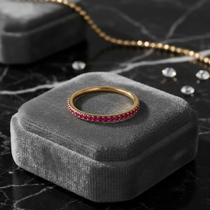 May include: A thin gold band ring featuring a continuous row of small, round, vibrant pinkish-red gemstones set along the outer circumference. The ring is displayed on a square, grey velvet-textured surface with rounded corners and a soft finish.