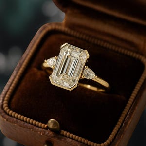 Puede incluir: Anillo de oro con un gran diamante central de talla esmeralda y dos diamantes más pequeños en forma de pera a cada lado. El anillo se presenta en un joyero de terciopelo marrón.