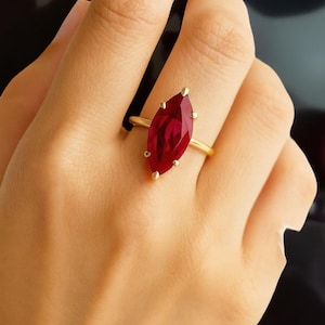 May include: A gold ring featuring a marquise-cut, deep red gemstone. The gemstone is held in place by four gold prongs. The ring's band is a simple, polished gold. The ring is shown on a person's finger.