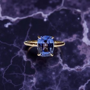 May include: A gold ring featuring a large, rectangular, blue gemstone. The gemstone is set in a four-prong setting, and the band is a simple, polished gold. The ring is displayed against a dark, marbled background.