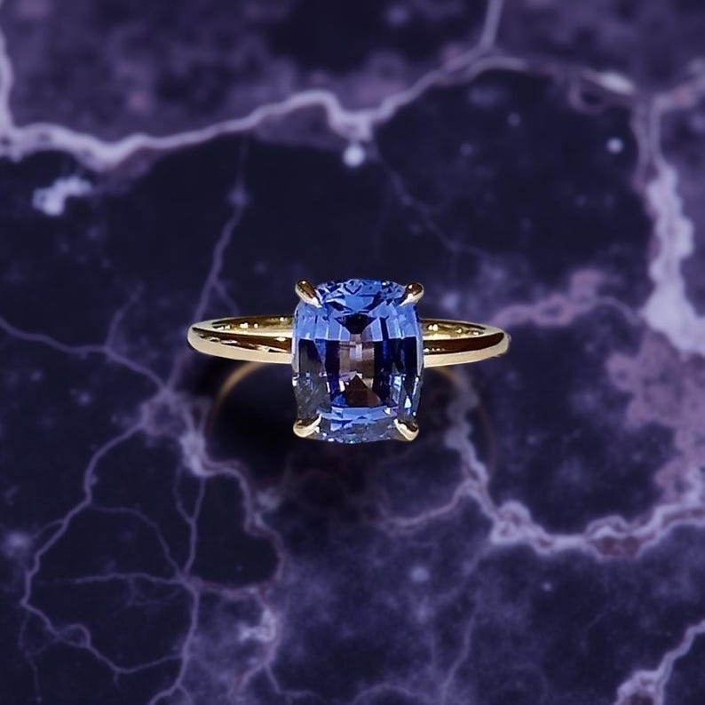May include: A gold ring featuring a large, rectangular, blue gemstone. The gemstone is set in a four-prong setting, and the band is a simple, polished gold. The ring is displayed against a dark, marbled background.