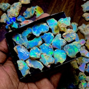 Peut inclure: Une collection d'opales brutes, présentant une iridescence vibrante de bleu, vert et jaune. Les opales sont disposées dans un plateau noir, avec d'autres pierres éparpillées autour. La texture rugueuse et le jeu de couleurs mettent en valeur la beauté naturelle des pierres précieuses.
