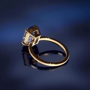 May include: A gold ring with a large, light blue gemstone. The ring's band is a simple, polished gold, and the gemstone is set in a four-prong setting. The ring is displayed on a reflective surface against a dark blue background.