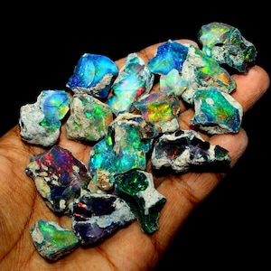 以下が含まれることがあります： 青、緑、赤、黄色の輝きなど、さまざまな色を示す、未加工のオパール原石のコレクション。 オパールは手で持たれており、暗い背景がその鮮やかで虹色の品質を強調しています。 これらの宝石は、自然な未切断の状態です。