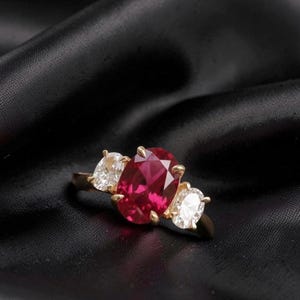 May include: A gold ring with a large, oval-cut ruby center stone and two smaller, pear-shaped diamonds on either side. The ring is set against a black background.