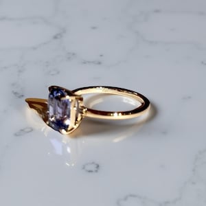 May include: A gold ring with a rectangular, light purple gemstone. The ring is set on a white marble surface. The band is thin and polished, and the gemstone is the focal point of the design.