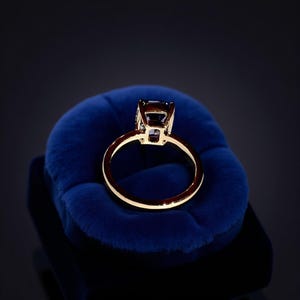 May include: A gold-colored ring with a large, square-cut, dark blue gemstone. The ring is set on a plush, dark blue velvet surface, creating a striking contrast. The band is thin and polished, highlighting the gemstone's brilliance.