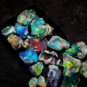Può includere: Una collezione di pietre preziose di opale grezzo in varie tonalità di blu, verde, rosso e nero. Gli opali sono esposti su una superficie nera.