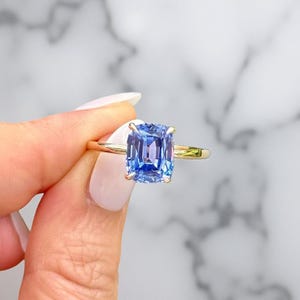 May include: A gold ring featuring a large, cushion-cut, light blue gemstone. The ring's band is a thin, polished gold. The gemstone is held in place by four prongs. The background is a blurred, marble-like pattern.