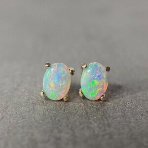 Puede incluir: Un par de pendientes de ópalo. Cada pendiente presenta una piedra preciosa de ópalo de forma ovalada con colores iridiscentes verdes, azules y amarillos. Los ópalos están engastados en un engaste de metal dorado.