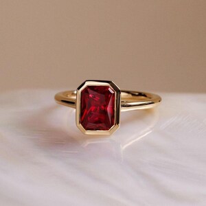Peut inclure: Une bague en or ornée d'une pierre précieuse rouge foncé de forme rectangulaire. La pierre est sertie dans une lunette en or, et le corps de la bague est en or uni. La bague est présentée sur une surface blanche texturée.