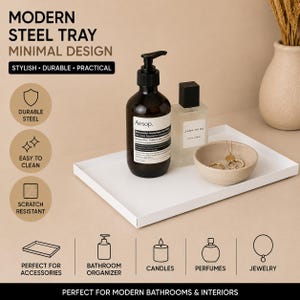 Può includere: Un vassoio rettangolare in acciaio bianco contenente un flacone a pompa marrone scuro con etichetta Aesop, un flacone di profumo in vetro trasparente con etichetta Zara Home e una piccola ciotola in ceramica beige con una collana e un anello.
