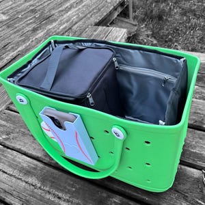 Puede incluir: Bolso tote verde brillante con interior negro y una bolsa de almuerzo aislada negra a juego. El bolso tiene un soporte para teléfono azul claro con un diseño de béisbol. El bolso tiene un asa y está sobre una superficie de madera.