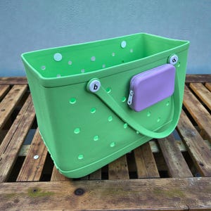 Puede incluir: Un bolso tote verde brillante con un asa a juego y una pequeña bolsa con cremallera de color lavanda adjunta. El bolso tiene forma rectangular con varios agujeros circulares pequeños y detalles blancos en forma de botón.