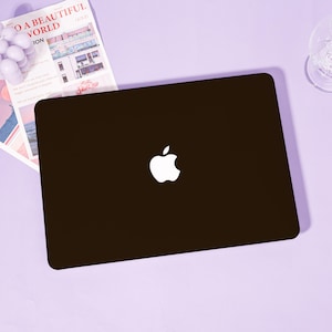 May include: A matte black laptop case featuring a centered white Apple logo. The rectangular cover has rounded corners and a smooth, solid dark finish. This protective shell is designed to fit securely over the exterior lid of a portable computer.
