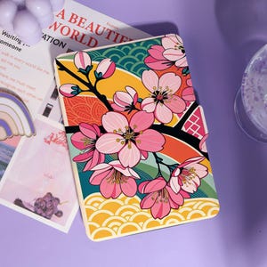 May include: A rectangular case with a vibrant floral design. The case features pink cherry blossoms on a branch against a backdrop of colorful stripes and patterns. The text "A BEAUTIFUL WORLD" is partially visible.