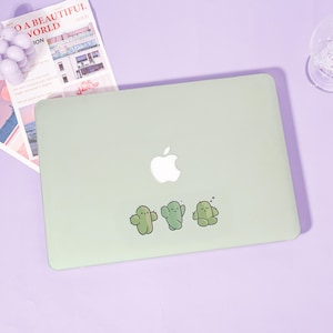 Peut inclure: Un ordinateur portable vert clair avec un logo Apple blanc et trois autocollants de cactus de dessin animé. Les autocollants sont verts avec des visages souriants. Un magazine avec le texte "TO A BEAUTIFUL WORLD" est en arrière-plan.